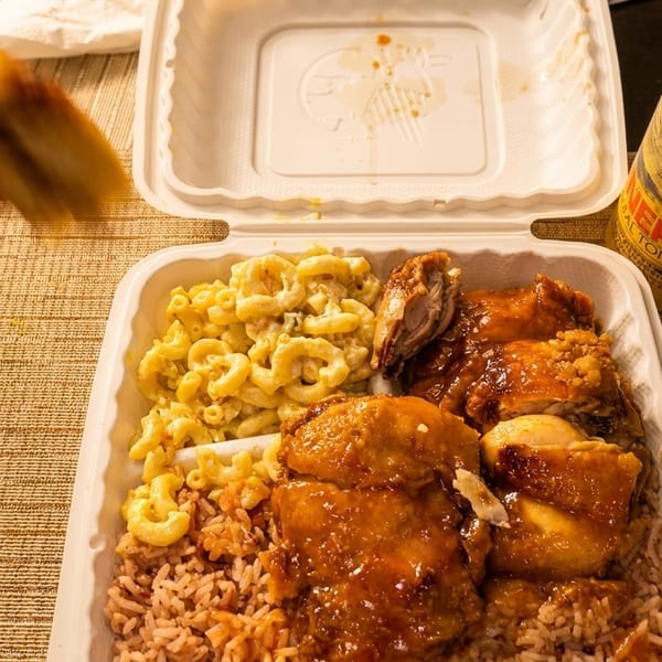 Takeout container with brown stew chicken, rice and peas, macaroni salad, and a drink.