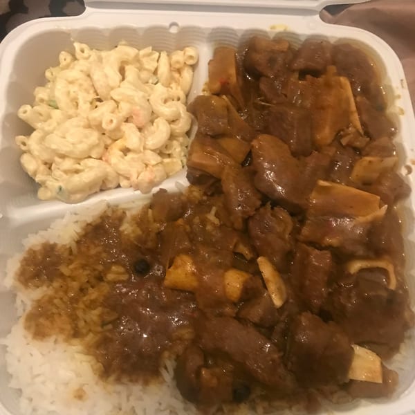 A takeout container holds macaroni salad, white rice with gravy, and a stewed meat dish.