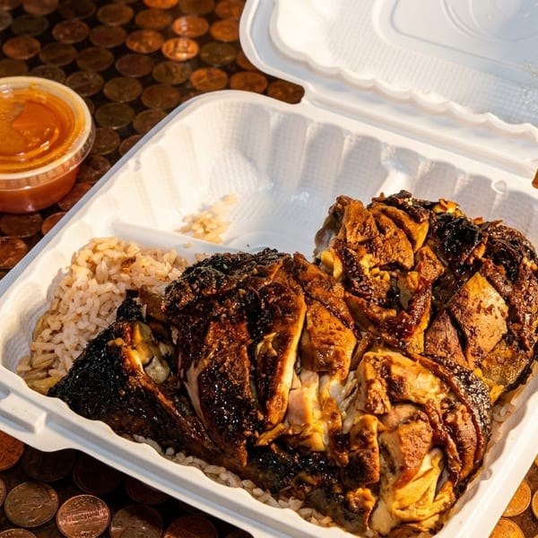 Jerk chicken and rice in a takeout container with two sauce cups on a penny-covered surface.